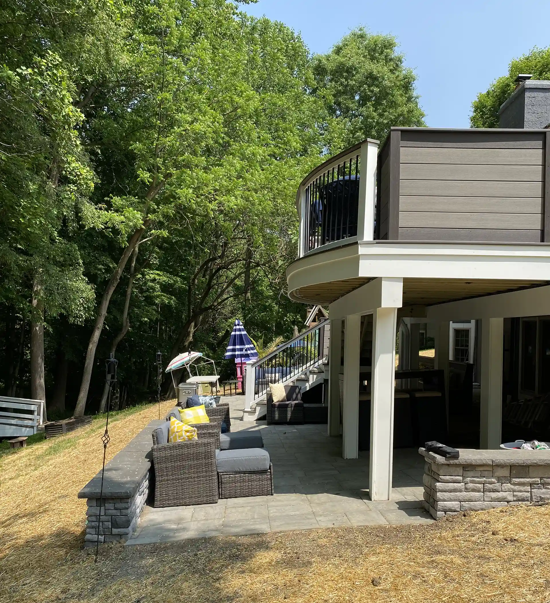 A curved deck with dark brown railings, supported by white pillars, overlooking a stone patio with outdoor furniture set against a backdrop of trees.