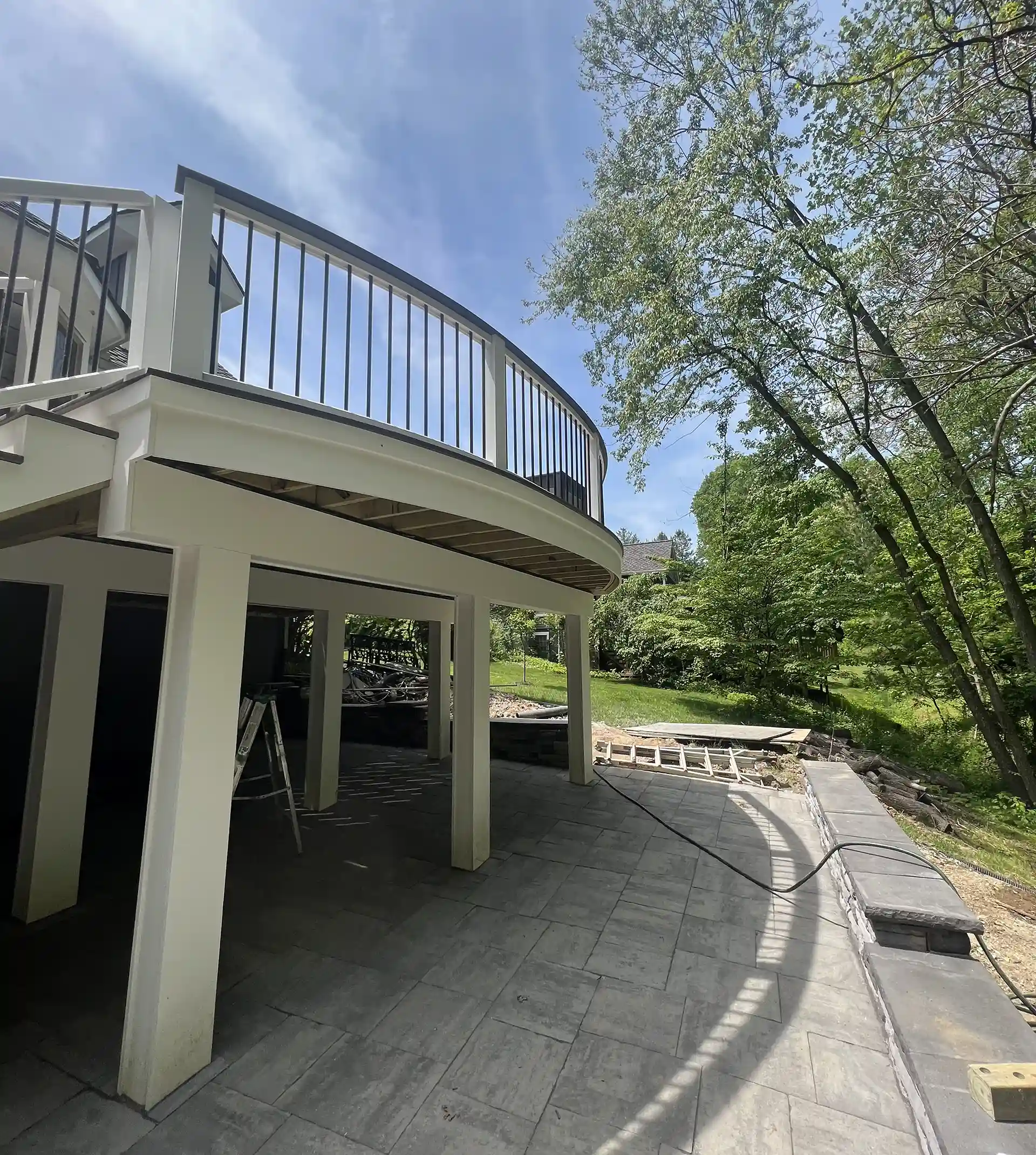 A curved deck with a white frame and black railings, elevated above the ground and supported by tall pillars, with a landscaped yard and stone patio visible below.