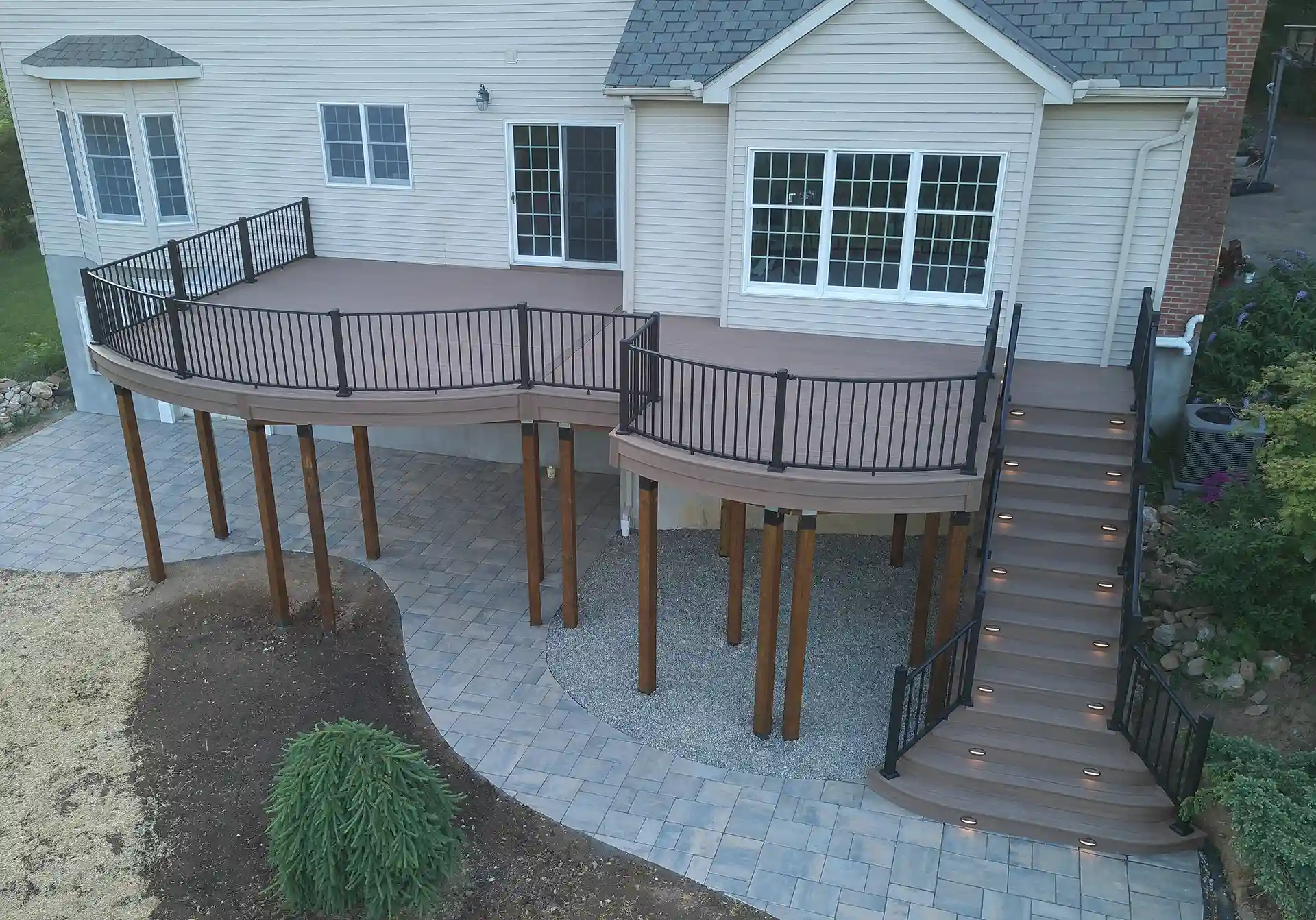 A large curved deck with black railings and lighting, elevated on stilts with a staircase leading down to a paved outdoor area, attached to a house with multiple windows.