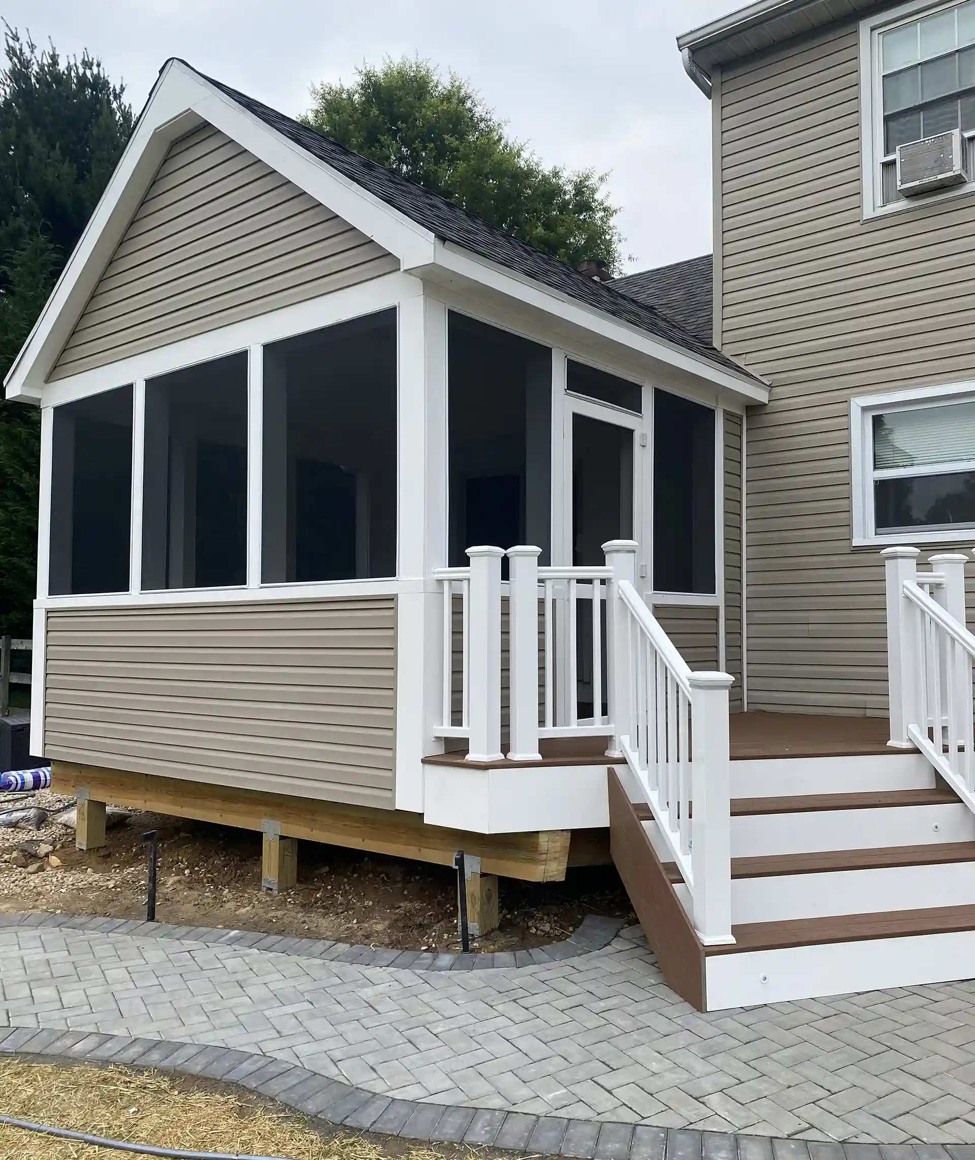 Backyard view of a sunroom with a raised deck and a wooden porch swing on a paver patio under mature trees.
