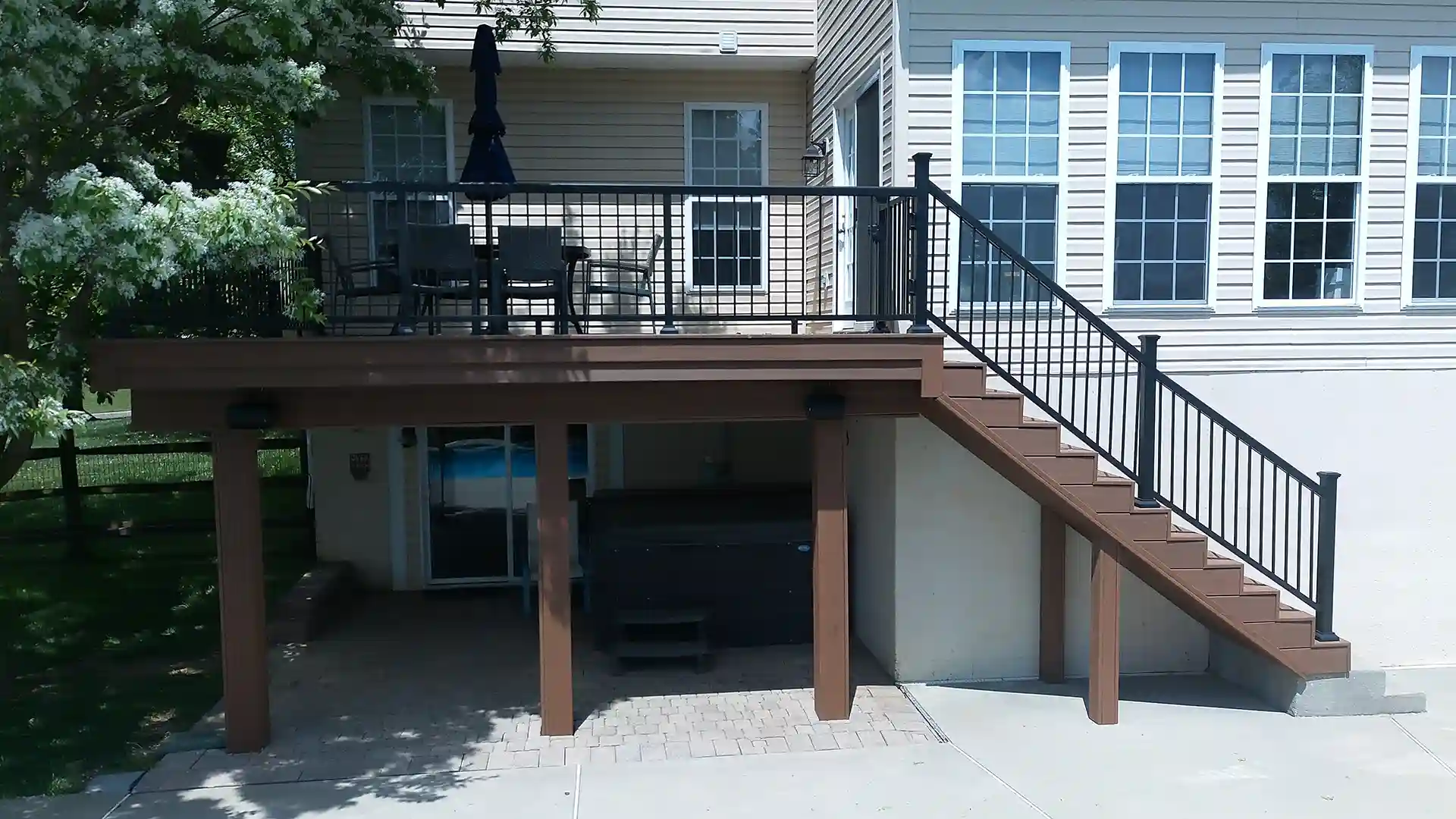Raised composite deck with black railings and stairs, creating a covered patio area below with pavers and a built-in hot tub.