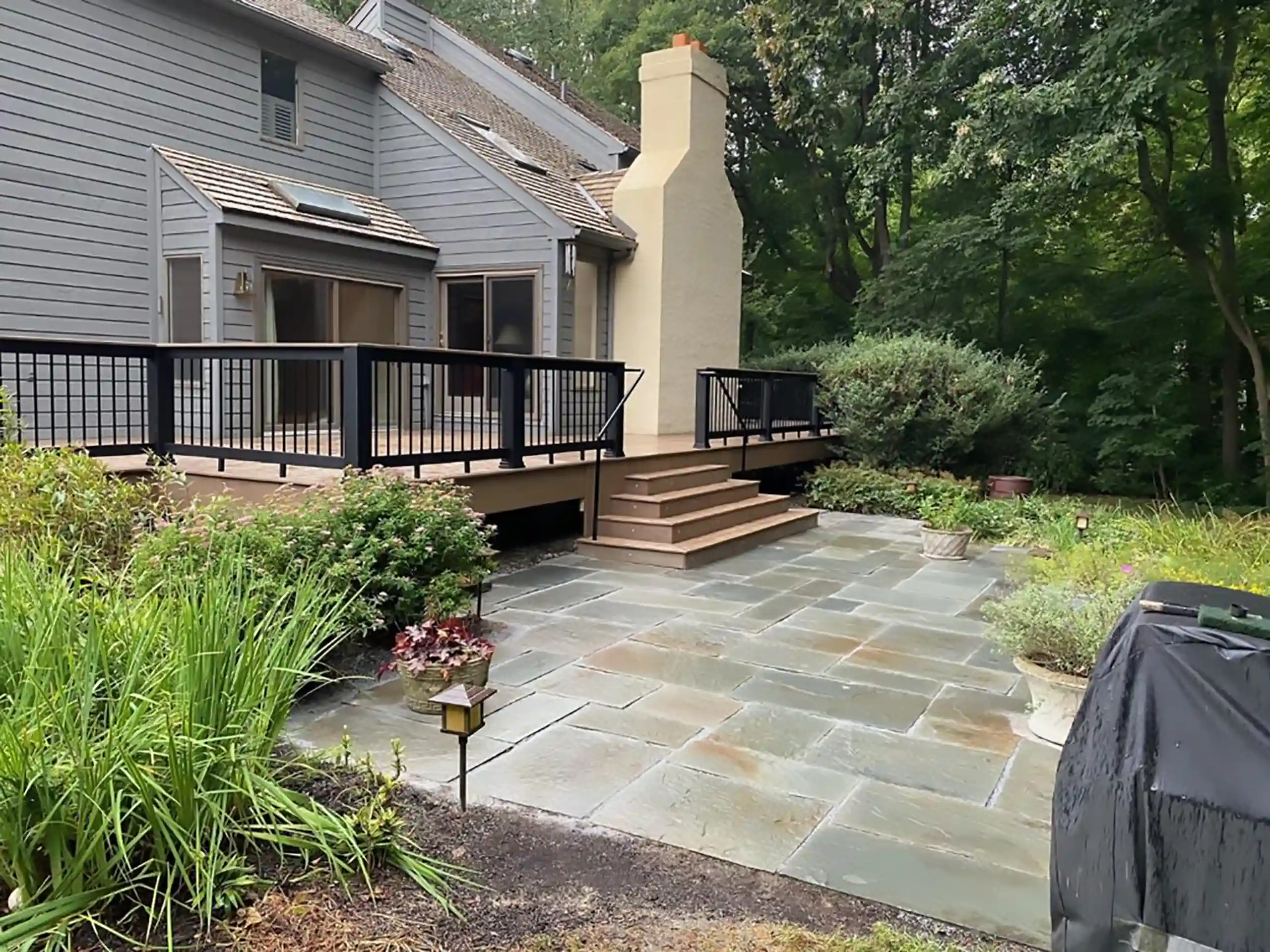 Deck with black railings and steps leading down to a stone patio, surrounded by lush greenery and garden plants.