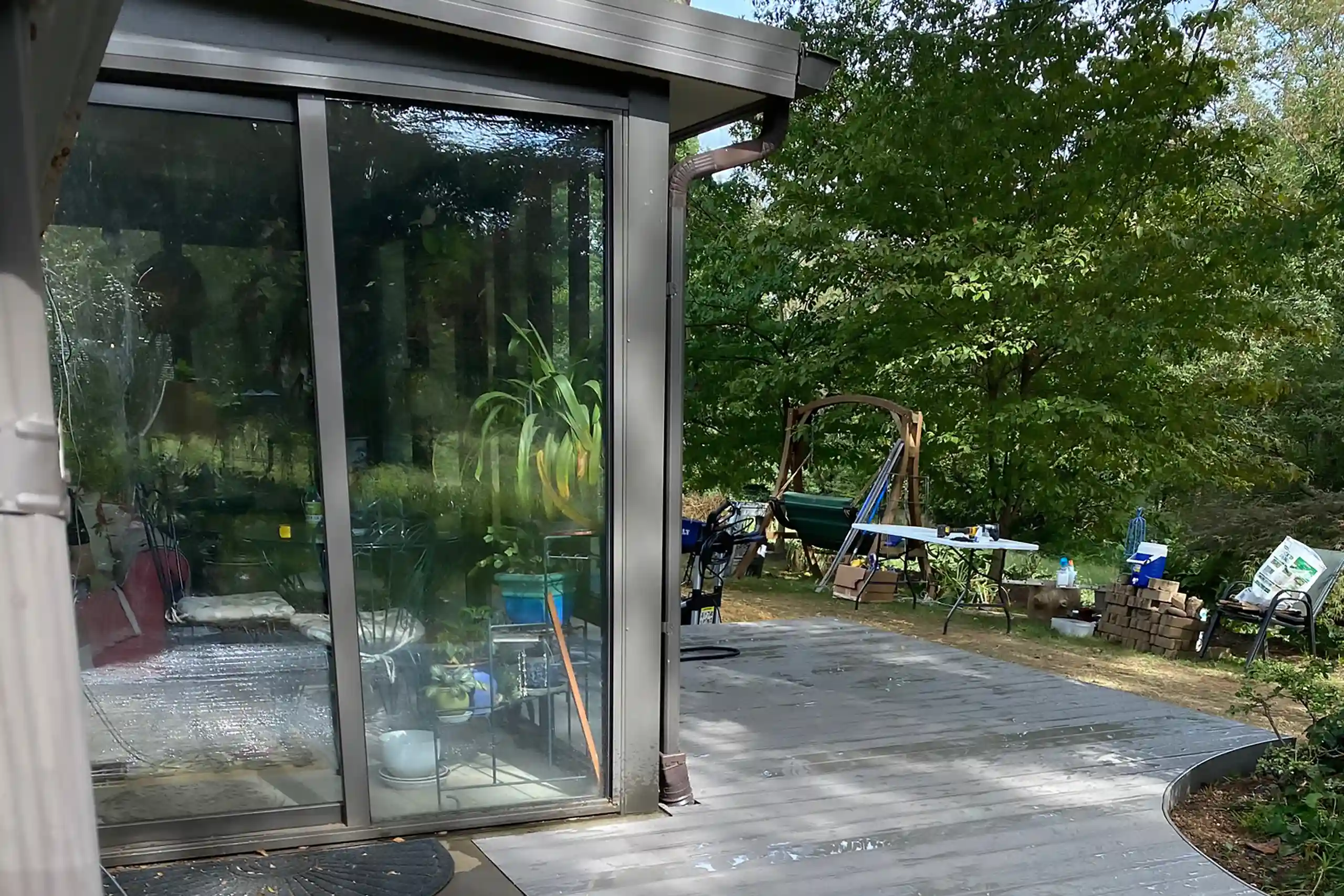 Close-up view of a gray composite deck connected to a sunroom with large glass windows, overlooking a wooded backyard with outdoor seating.