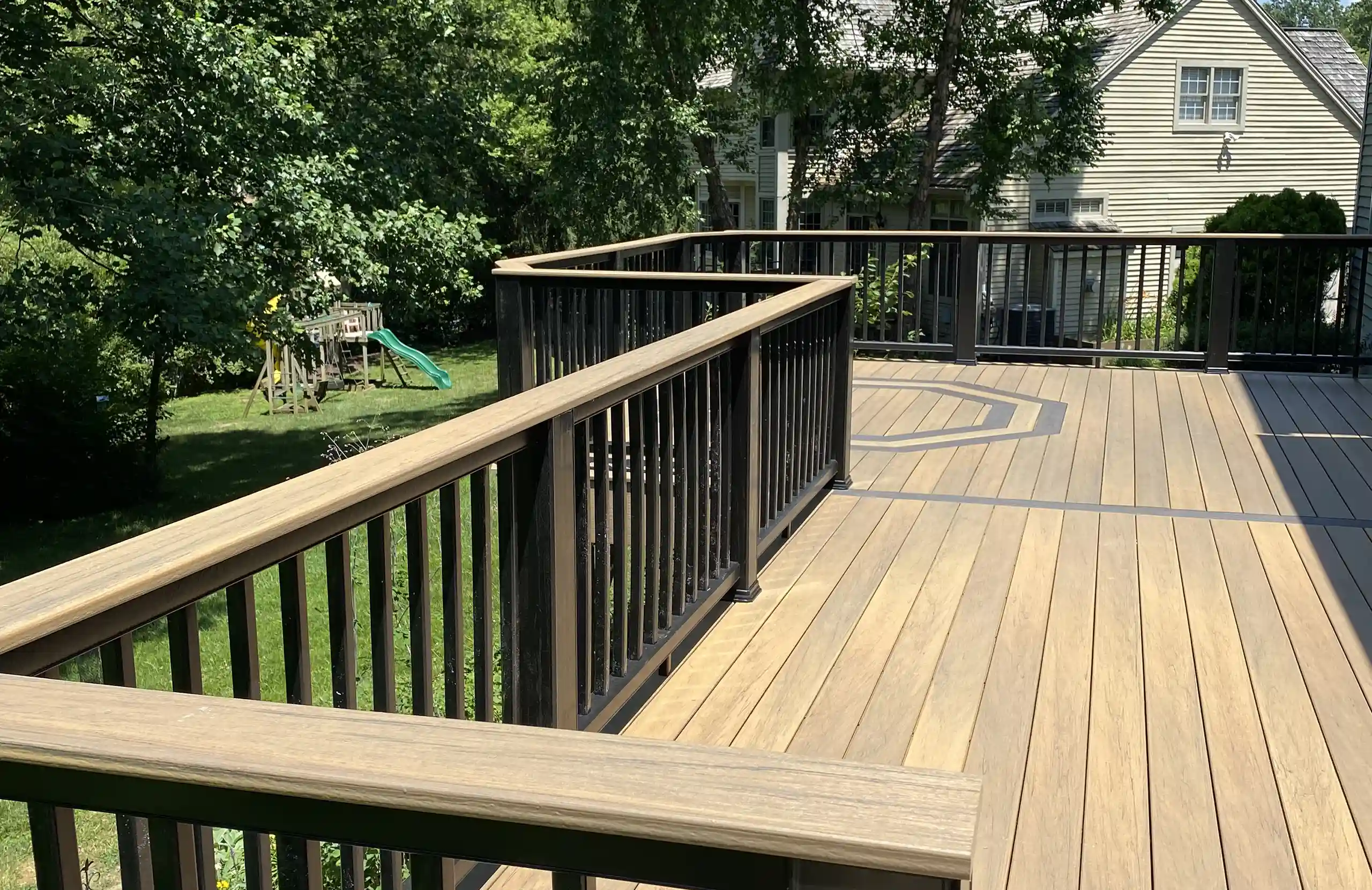 A close-up view of a wooden deck with black railings, featuring a circular design in the decking pattern, surrounded by greenery and a residential house in the background.