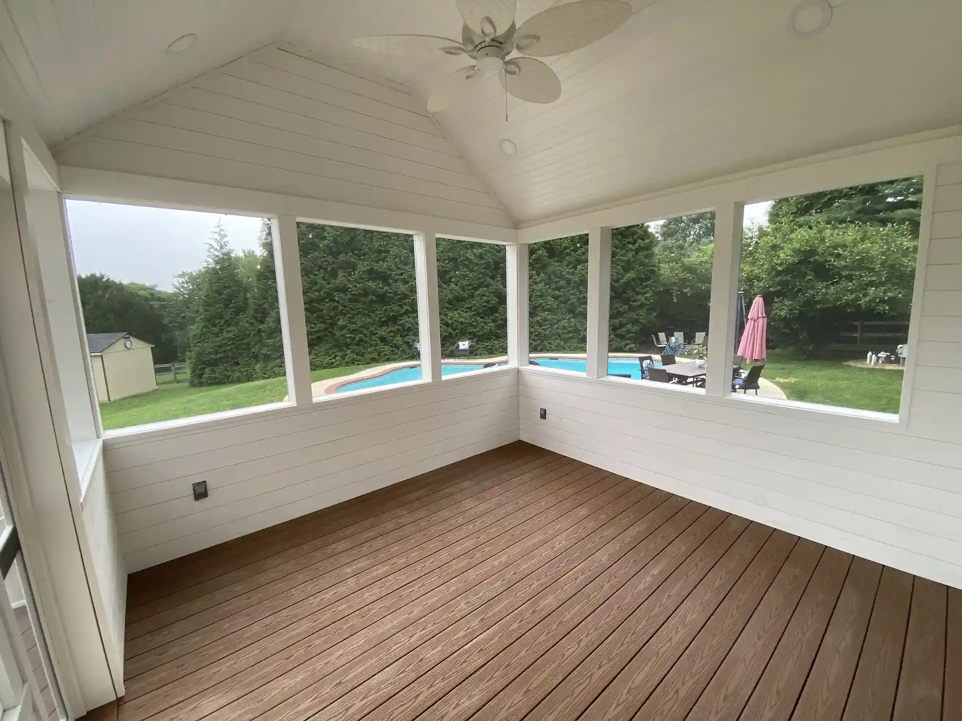 Interior view of a white screened-in sunroom with a vaulted ceiling, ceiling fan, and brown composite flooring, overlooking a backyard pool and patio.