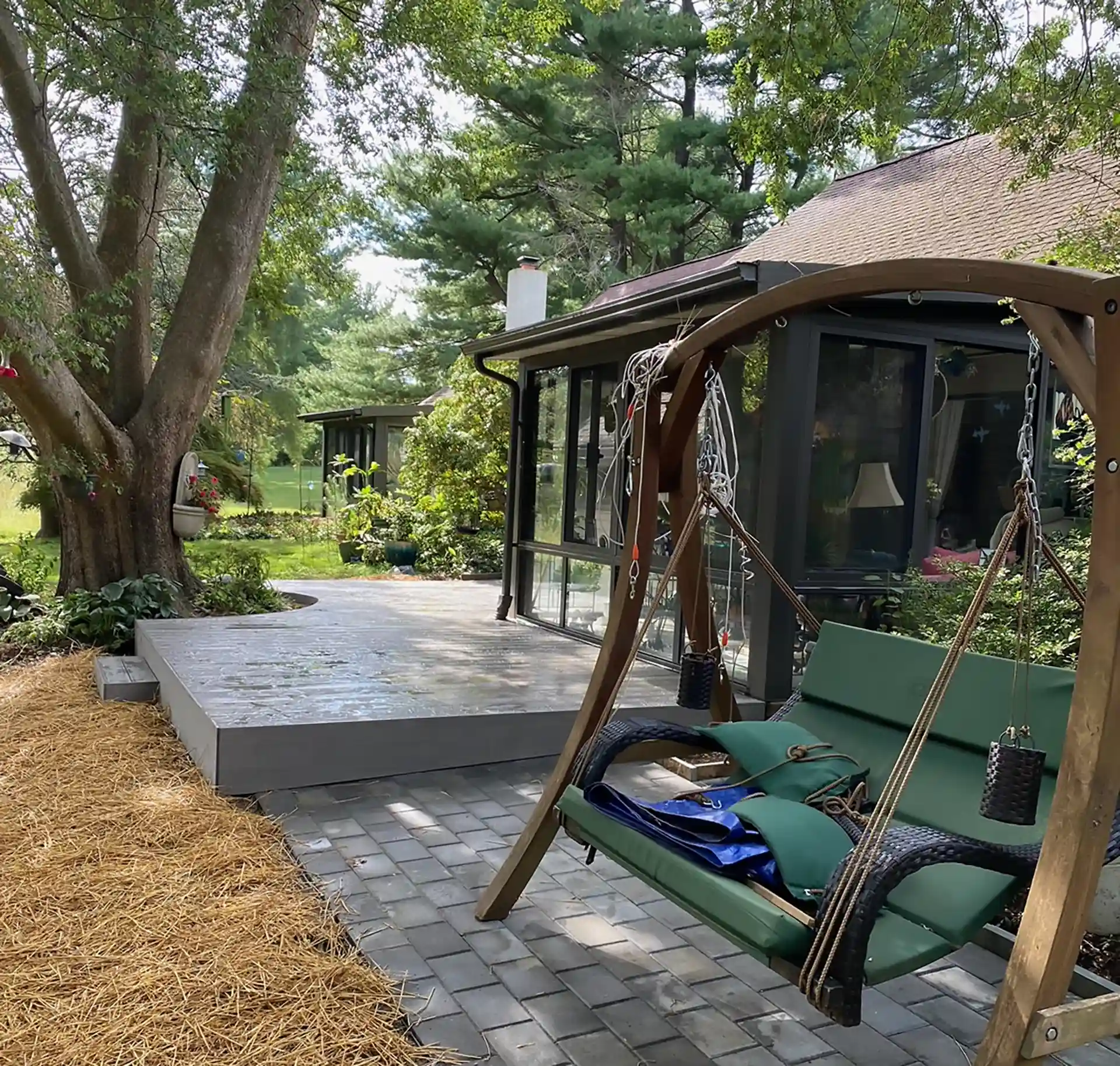 Backyard view of a sunroom with a raised deck and a wooden porch swing on a paver patio under mature trees.