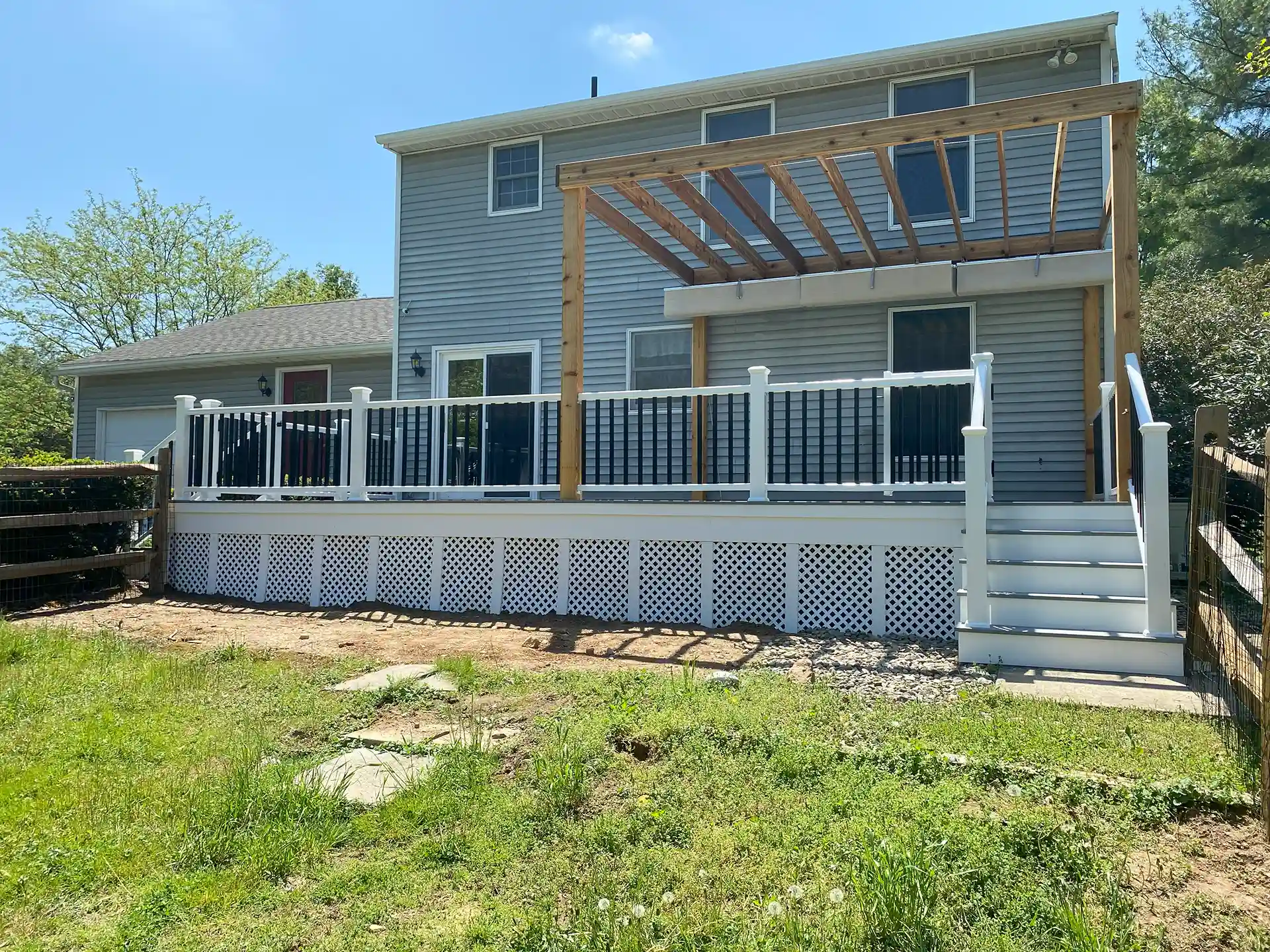 Backyard deck attached to a two-story house with white lattice deck skirting, white railing with black balusters, wooden pergola overhead, and stairs leading to the yard. 