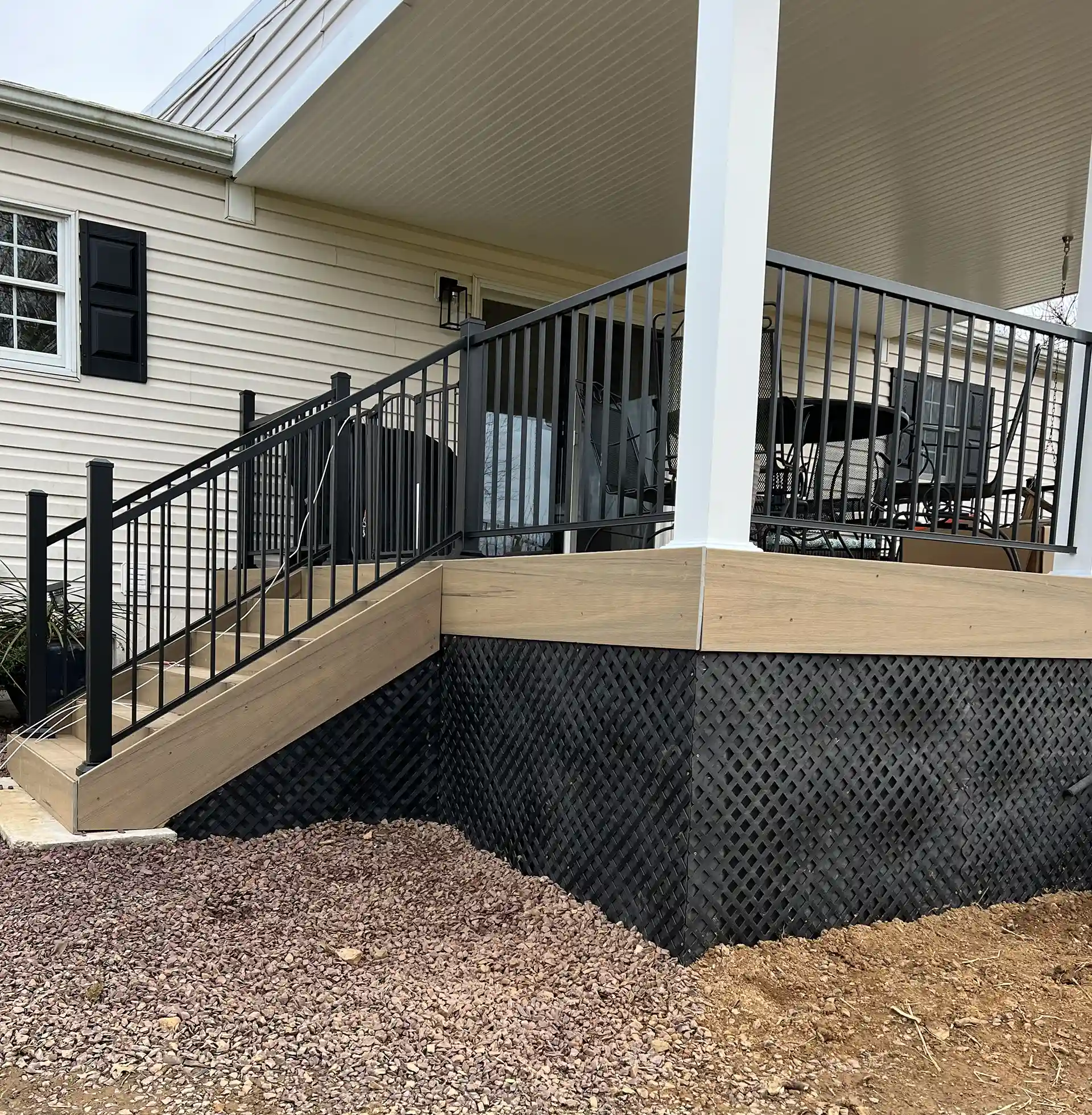 Corner of a deck with black metal railing, light wood decking skirting, black lattice skirting below, and dirt and gravel ground around the deck. 
