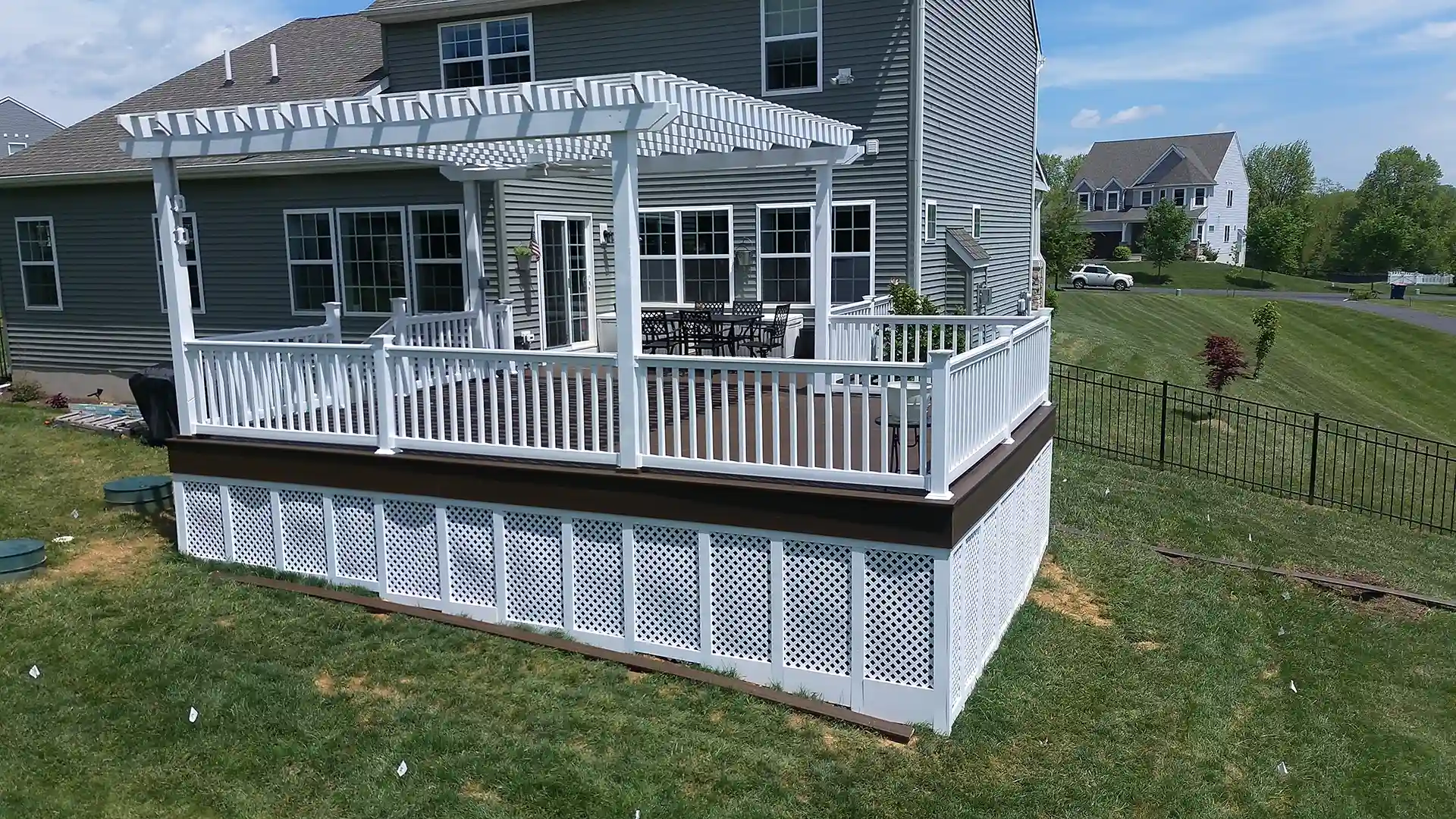 Deck attached to a house with white railing and white lattice skirting, pergola overhead, and grass lawn surrounding the deck. 