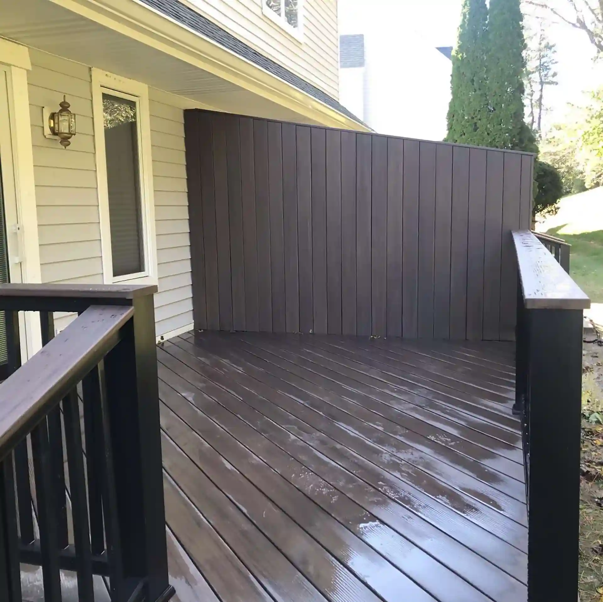 A backyard deck featuring a vertical privacy wall made of dark brown wood panels. The deck has a wooden floor and a black railing. 