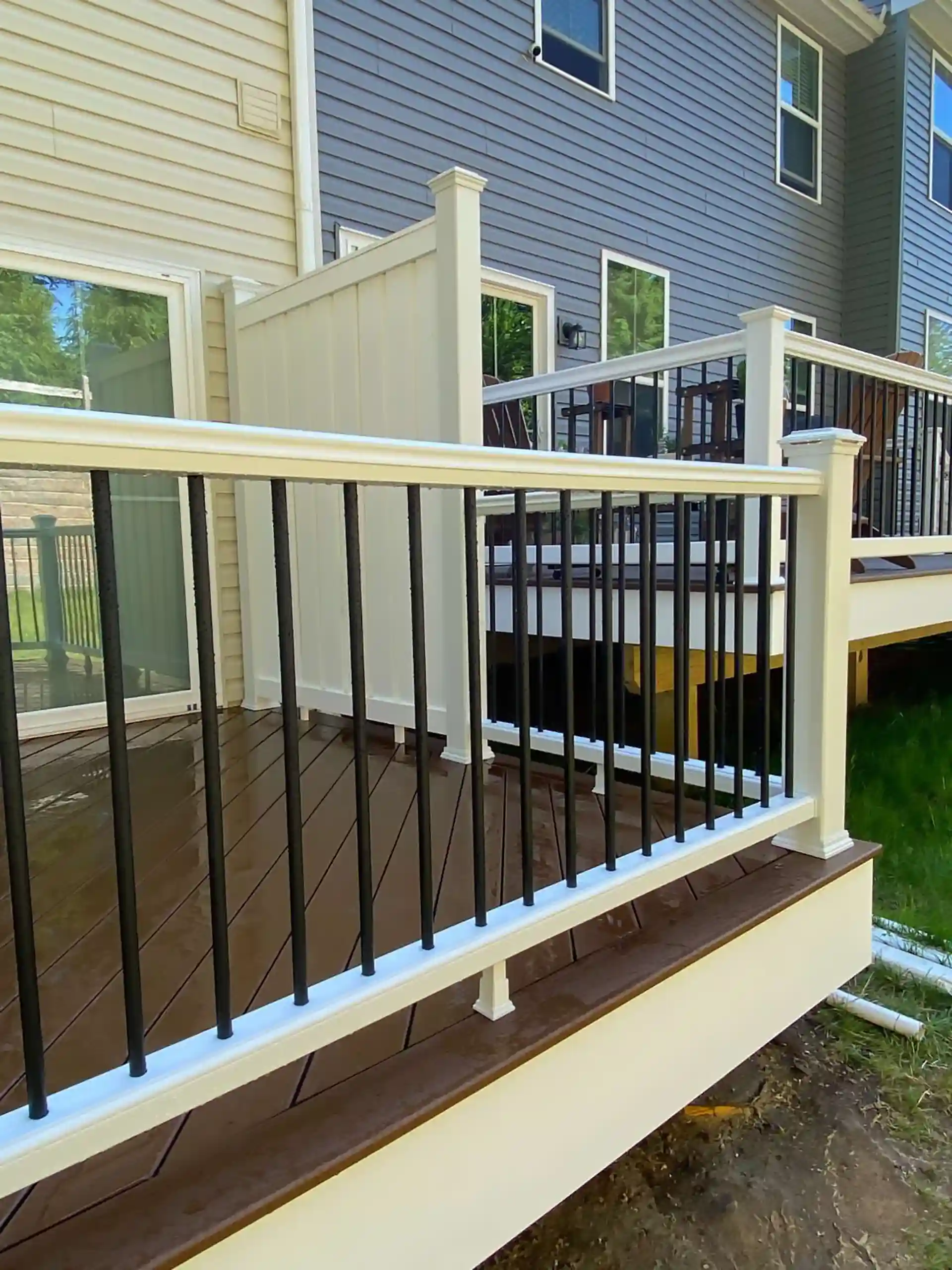 A backyard deck with a white vinyl privacy wall and a black metal railing with vertical balusters. The deck has a wooden floor, and the background shows a multi-level home. 