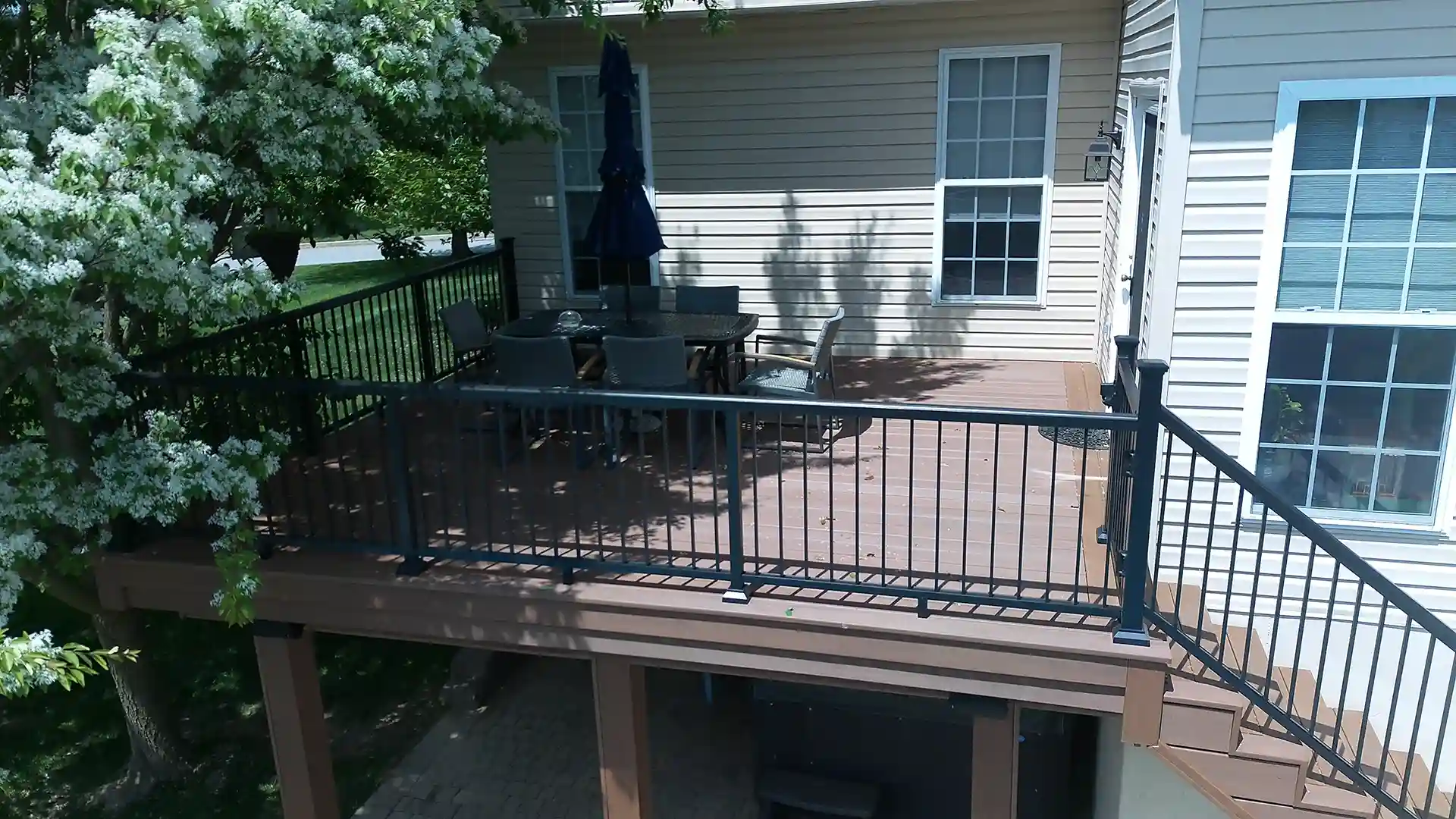 Elevated brown composite deck with black metal railing, outdoor dining set, and stairs leading down, surrounded by greenery and house siding. 