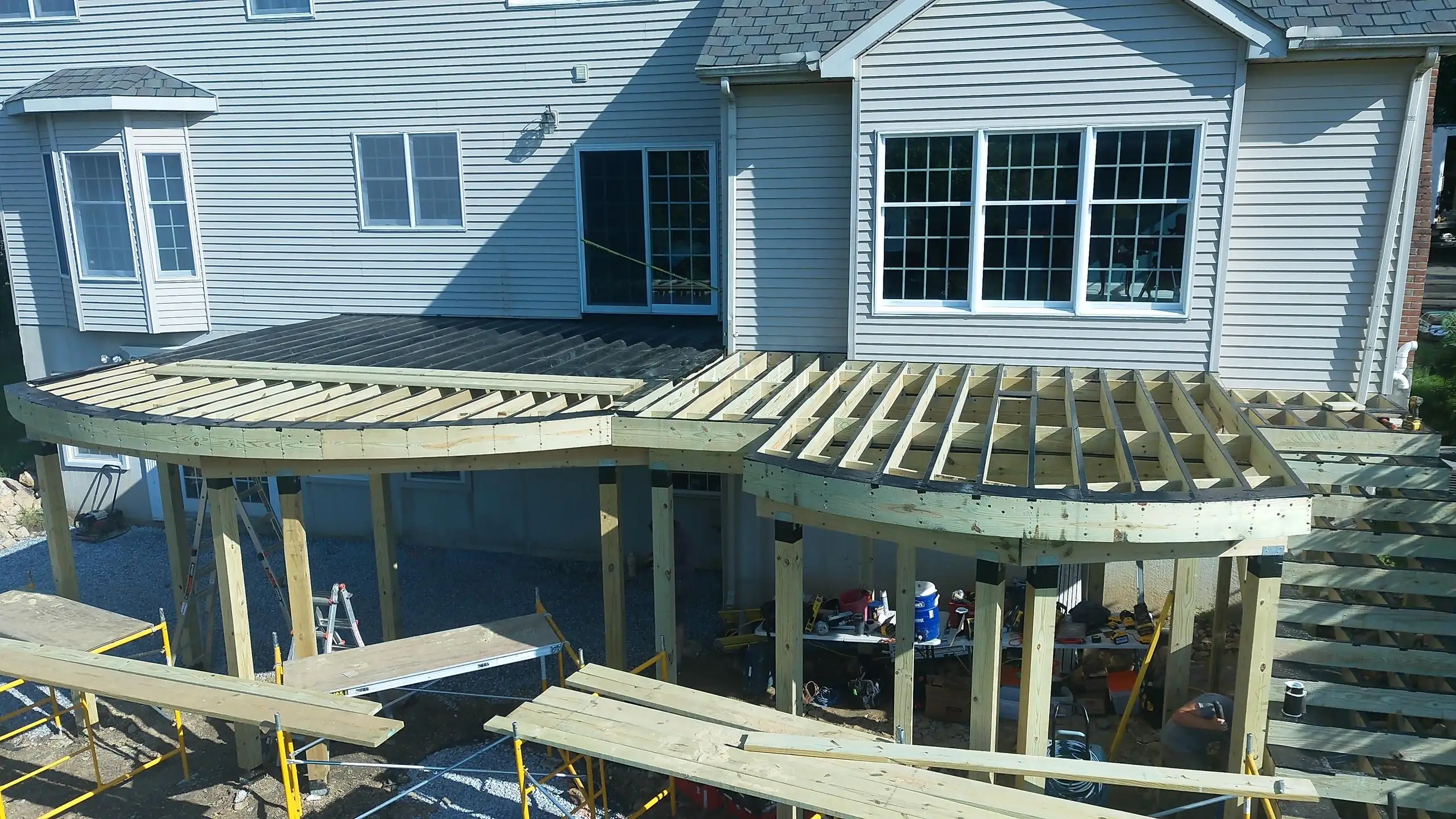 how to frame a deck High-angle photo of a multi-level wooden deck under construction at the back of a light gray house. The framing shows curved edges, and some dark deck boards are visible on the upper level.