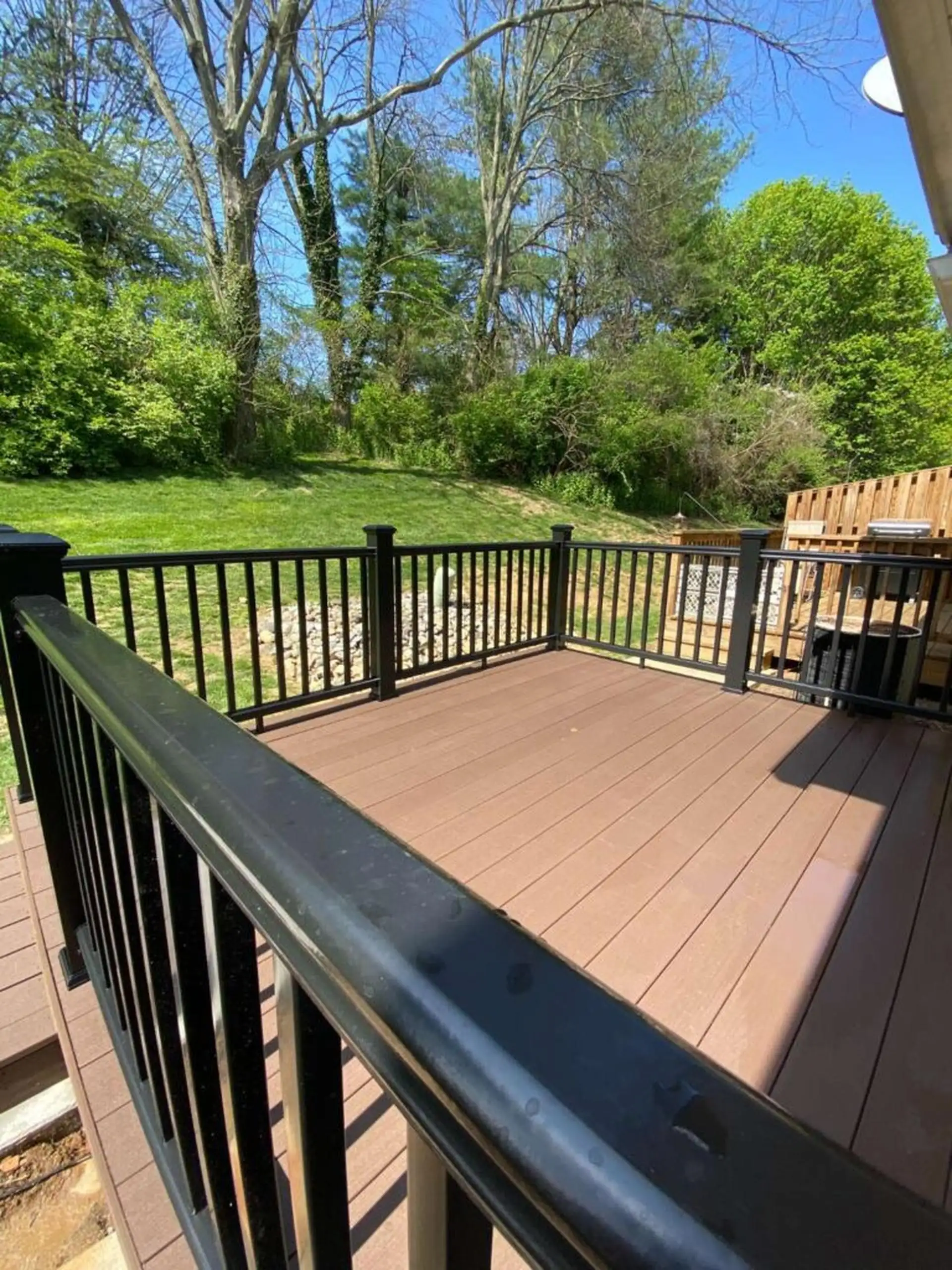 composite deck cost per square foot An outdoor deck with composite, brown decking material and black railings, overlooking a grassy, wooded backyard area on a sunny day.
