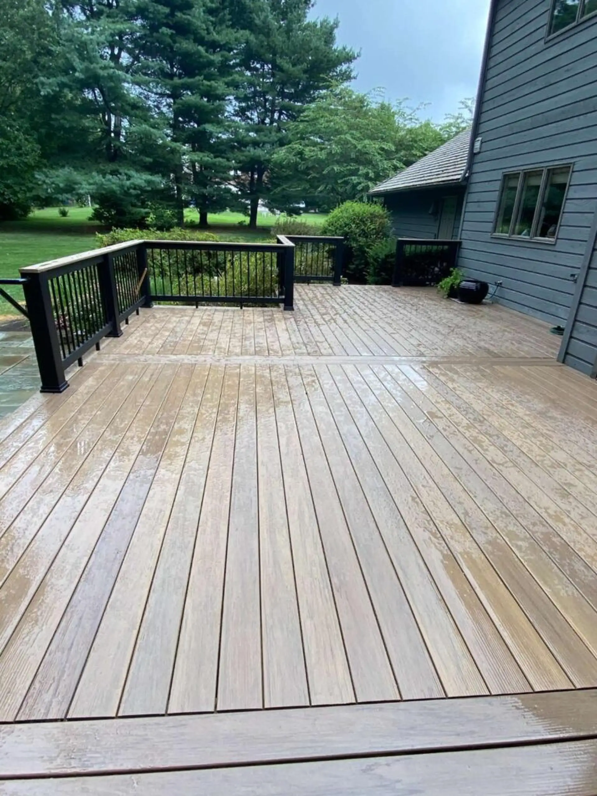 composite deck replacement cost A wet, composite deck in a brown-gray color, with a black metal railing overlooking a lush, green yard and surrounding trees. The deck connects to a dark-gray or blue-gray house on the right.