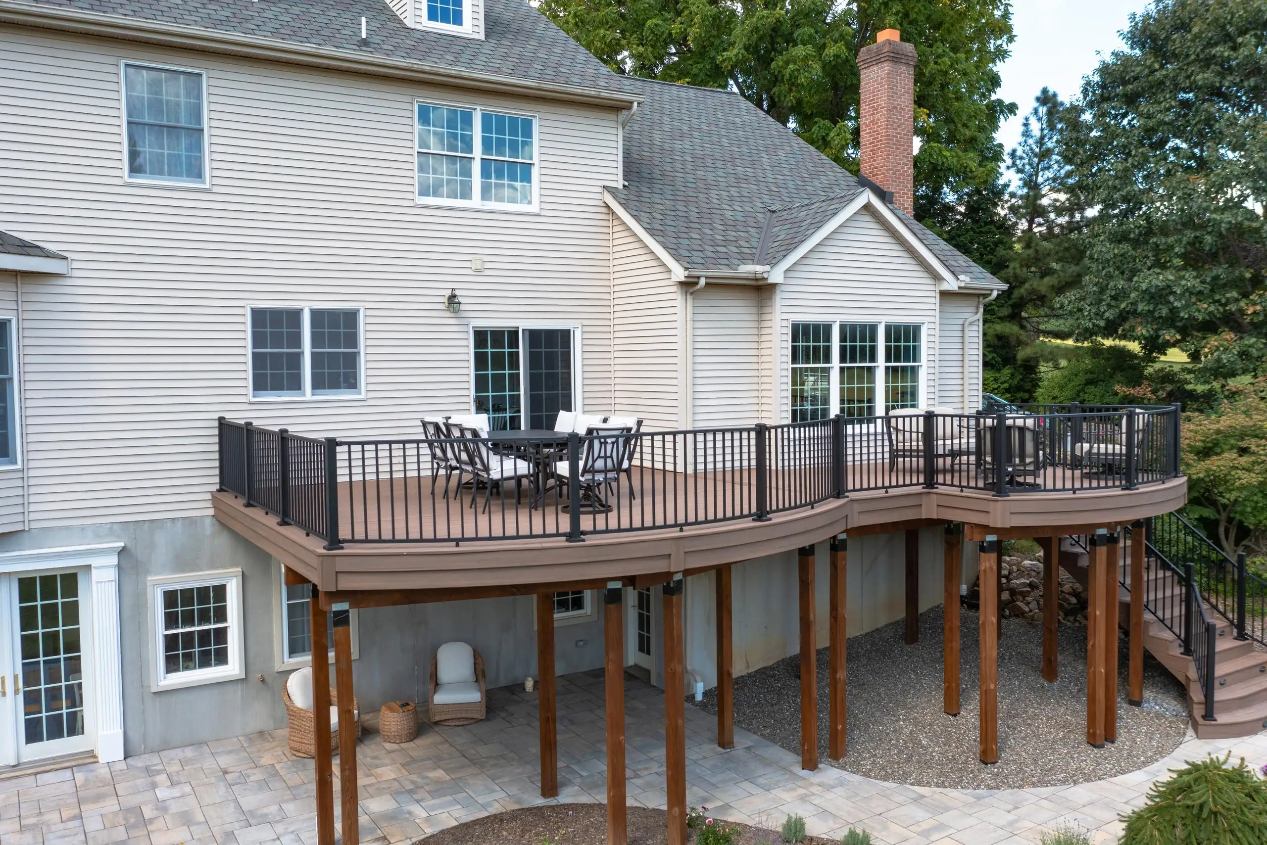 does building a deck require a permit Large, multi-level composite deck with curved edges, attached to a beige house. It features dark brown boards, black metal railings, and supports over a patio and gravel area.