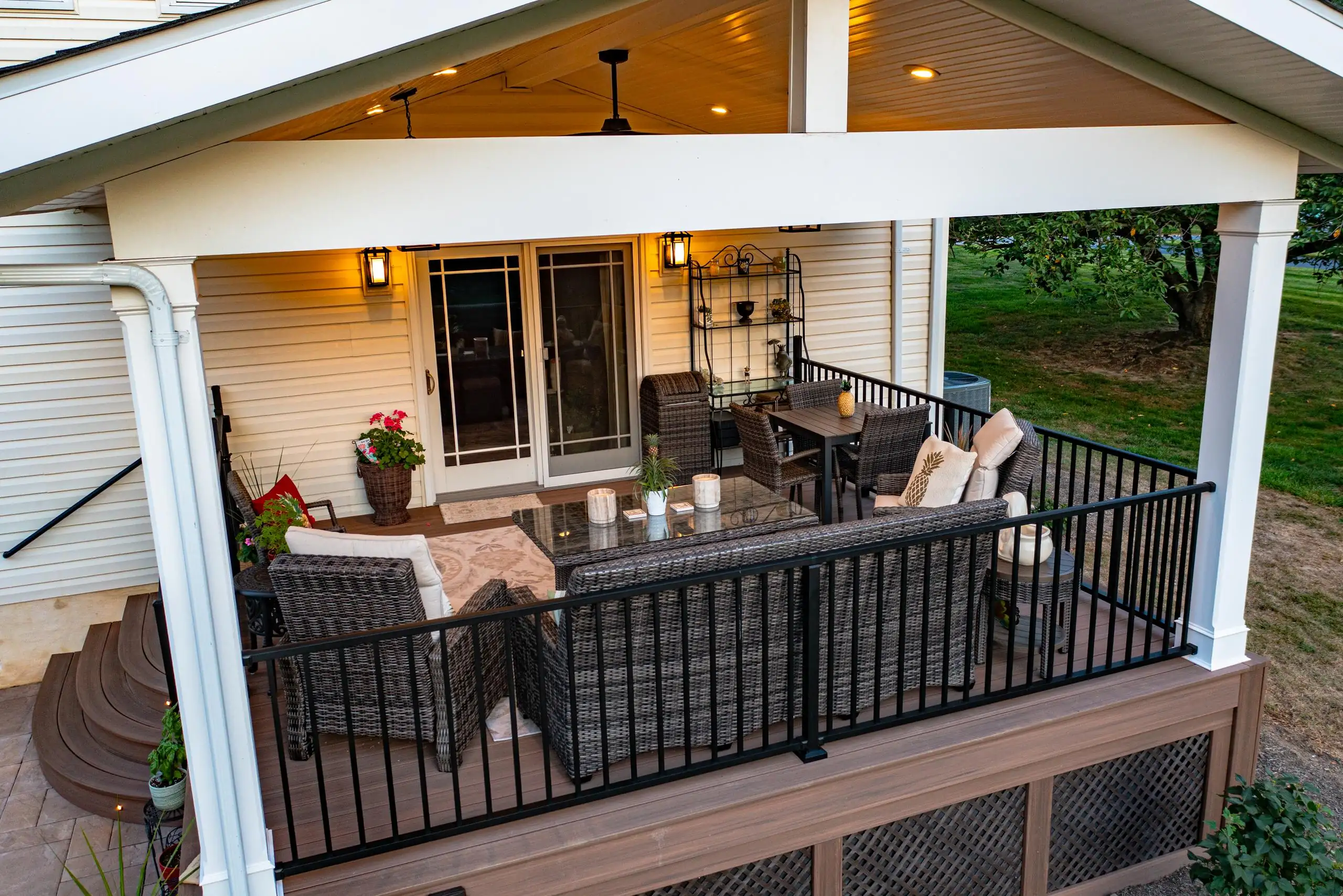 when a deck permit is mandatory A covered outdoor deck featuring dark brown wicker furniture, including a sofa, chairs, and a table, arranged for seating and dining. The deck has a light brown composite floor and is enclosed by black metal railings and white posts. The ceiling has exposed white beams, recessed lighting, and a ceiling fan.