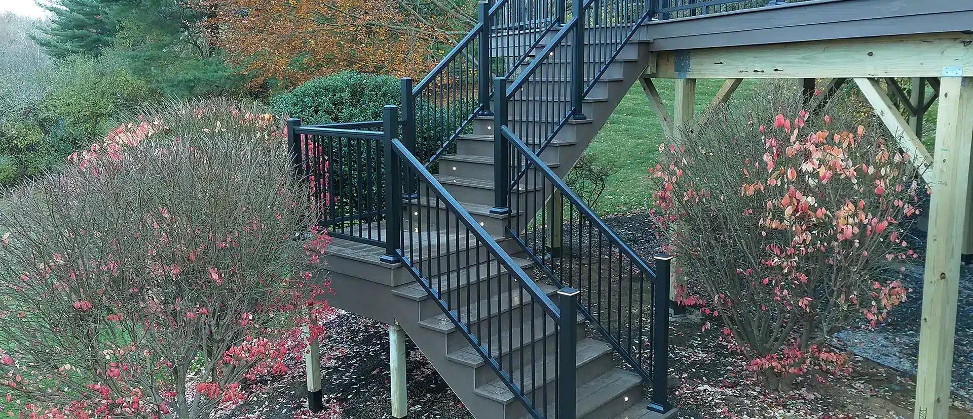 A set of outdoor stairs with brown composite decking and black metal railings descends from an upper deck area into a yard with bushes featuring red and green foliage. Small lights are embedded in the stair risers.